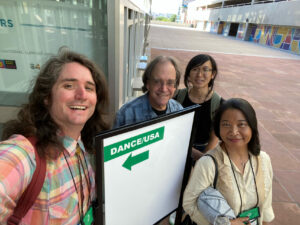 Four people stand outdoors smiling by a sign that says "Dance/USA" with an arrow.