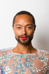 A non-binary person, wearing a sparkly top and bright lipstick.