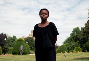 A Black woman, wearing a black dress, stands in front of a lawn with trees.