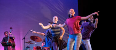 Three dancers smile and point different directions in the foreground. Behind them a jazz band plays.