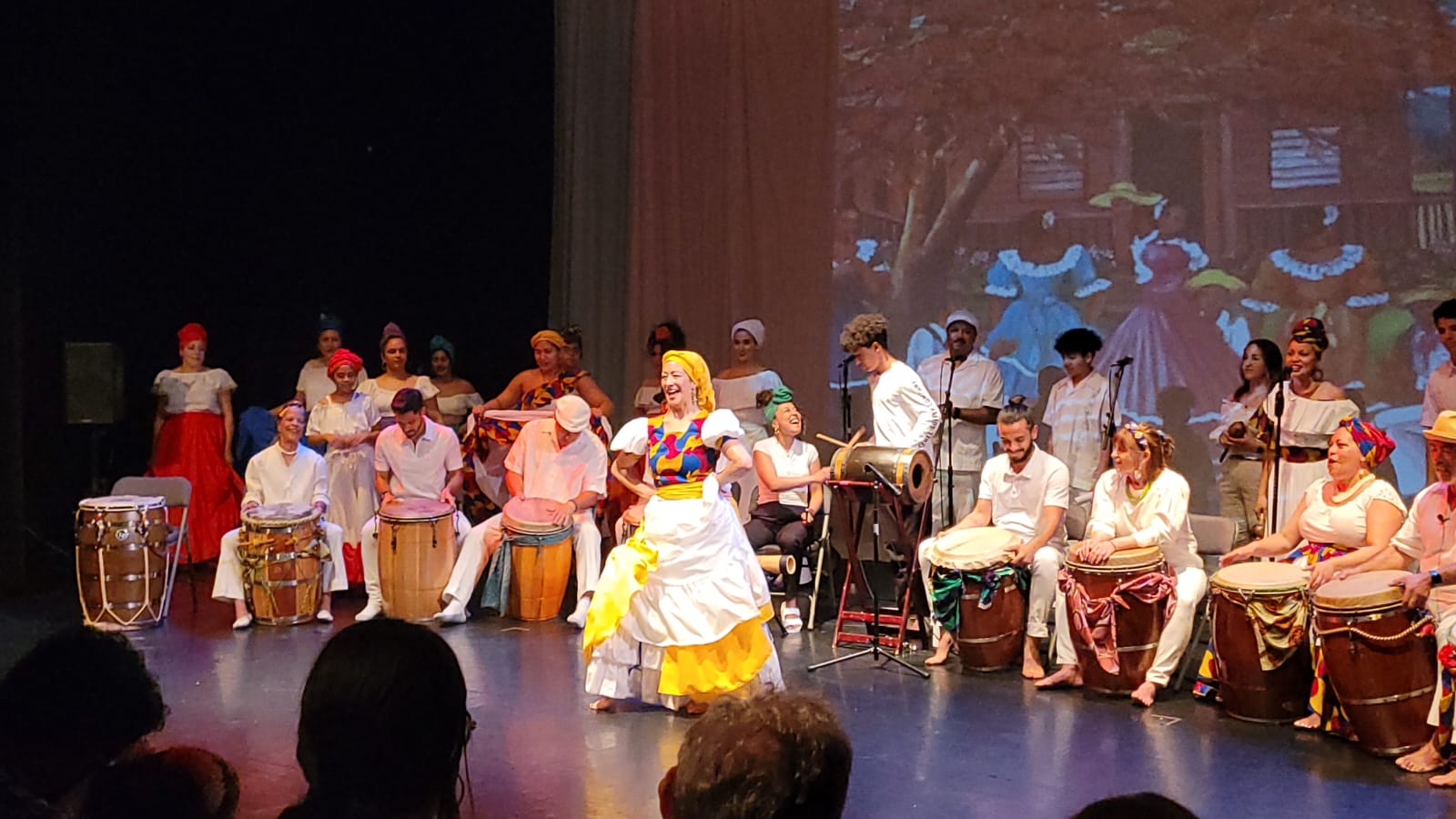 A large band of drummers and other instrumentalists behind a dancer who smiles on stage