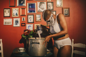 A Black performer in white underwear and a white beaded face covering pours salt into a large pot. The wall behind them is red and has lots of framed photos of people.