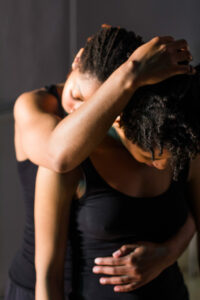 Two Black dancers wearing all black. One stands behind the other and holds them with a hand on their stomach and the other on their head.