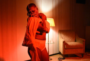 A performer lit in very red lighting wearing underwear, draping fabrics, and a headpiece over their face with their arms wrapped around themself.