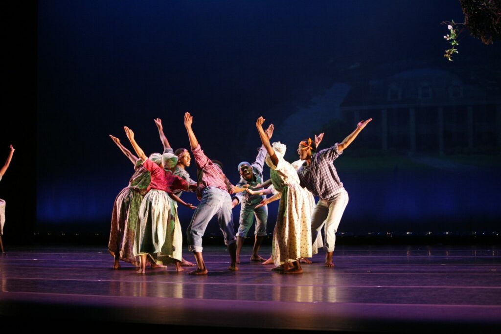 A group of dancers onstage in a circle facing each other with one arm reaching to the center of the circle and one arm extended behind them. They appear to be shouting. 