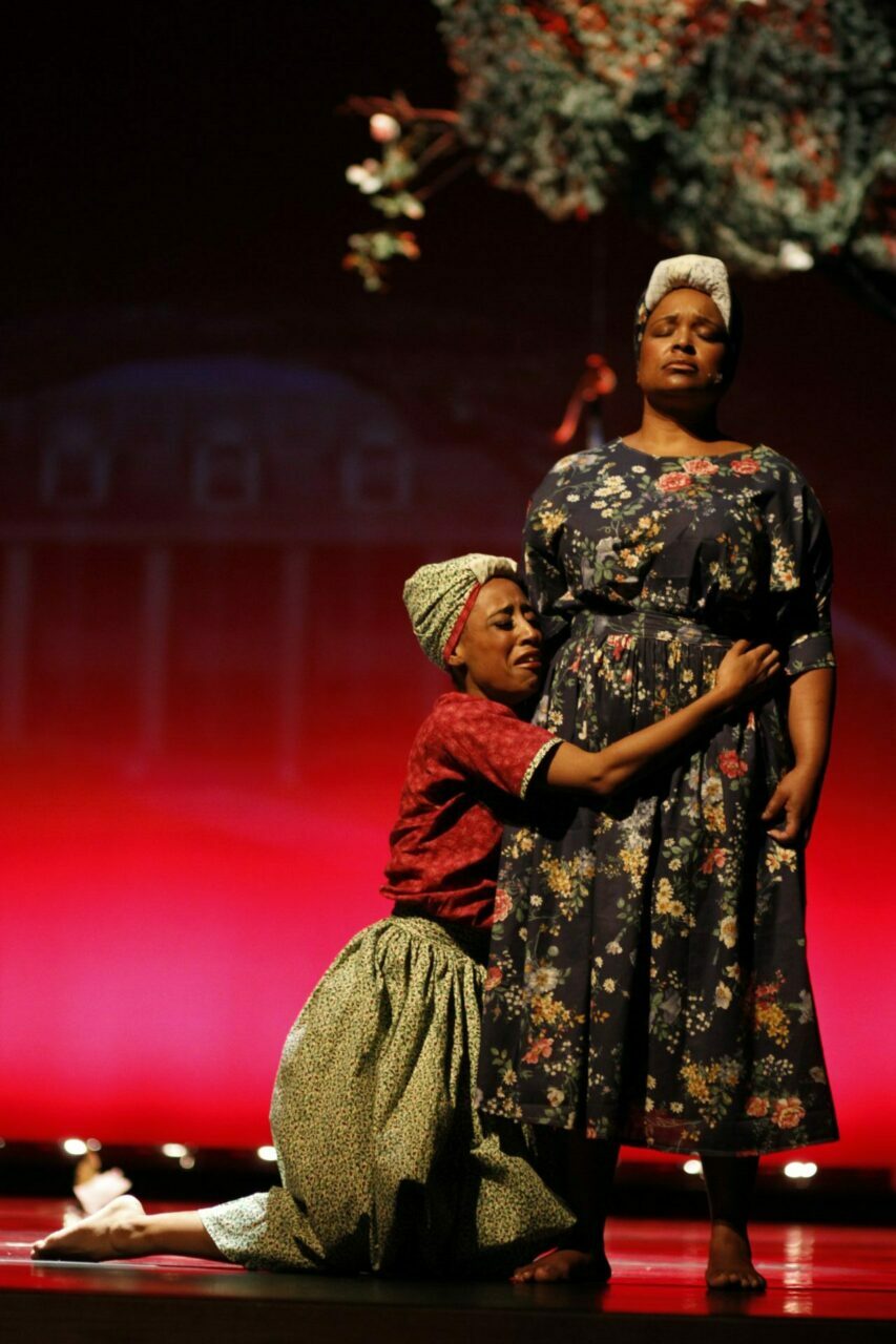 A Black dancer kneeling and wrapping their arms around a standing Black dancer. They both have pained expressions on their faces and are wearing head wraps and long skirts. 