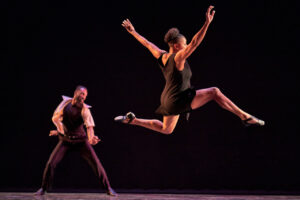 A dancer jumps high in the air with one leg bent in the front and the other bent in the back, throwing their arms up high. Another dancer in the background stands on the floor and is grooving.