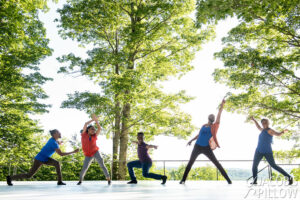 Five Black dancers all strike poses and smile broadly on an outdoor stage surrounded by green trees and foliage and a blue sky