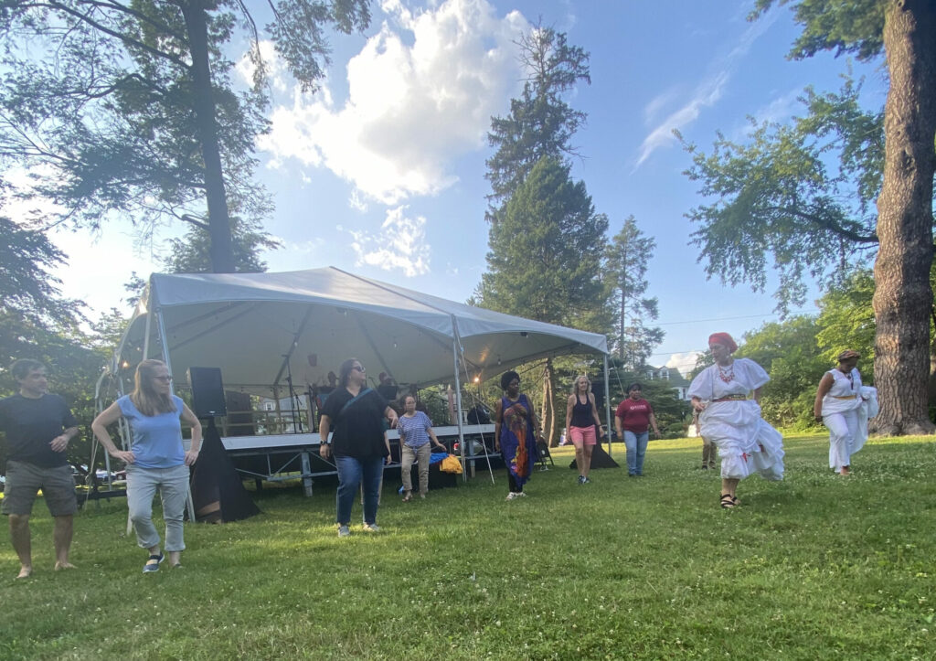 Standing on green grass with a bright blue sky behind them, dancers in white dresses show a diverse group of people movements