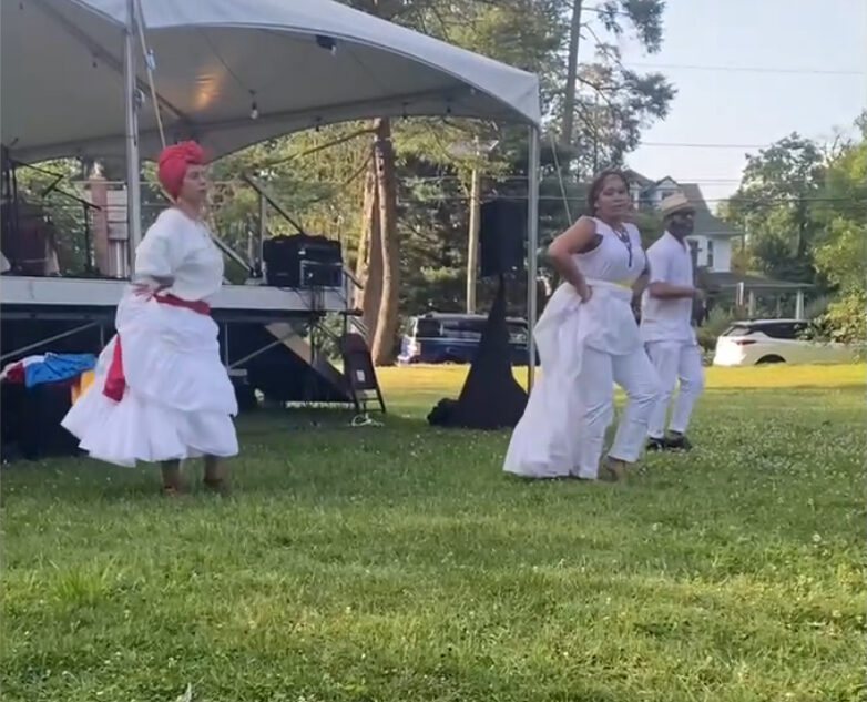 Three dancers wearing white dancing on green grass outdoors.