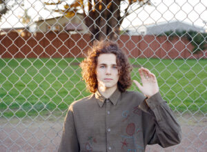 A person with curly dark hair standing outdoors in front of a chain link fence. They are wearing an embroidered button down shirt and waving their hand.