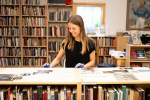 A white woman with long dirty blond hair wearing gloves and arranging papers on the top surface of a bookshelf. She is smiling and shelves of books surround her..