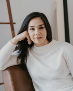 A woman with dark long hair wearing a white sweater smiling slightly and leaning her head into her hand.
