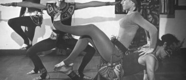 A black and white photograph of four dancers. In the background, a woman and man reach their arms out in squatting and perching postures. In the foreground, a woman site on a mans back and they touch feet. They all wear headresses and a mix of dance clothes and traditional garb.