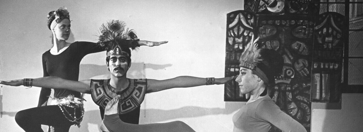 A black and white photograph of four dancers. In the background, a woman and man reach their arms out in squatting and perching postures. In the foreground, a woman site on a mans back and they touch feet. They all wear headresses and a mix of dance clothes and traditional garb.