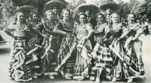 A black and white photo of a row of women wearing mexican folkloric long skirts. Behind them are men in large hats. All are smiling.