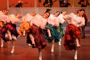 A group of women with white puffy shirts and colorful skirts dancing on stage. They kick their right legs up and lean to the left.