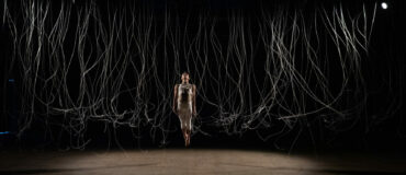 A performer jumping straight up in center stage with their body in a long straight position. All around them on the stage long silver tendrils fall from the ceiling.