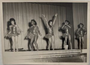 Black and white vintage photograph showing five children in shiny costumes dancing on stage. The center child, Dianne Walker, has her arms up in a "V" shape.