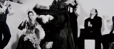 A black and white image of a flamenco dancer dancing with a group of four singers and musicians behind her