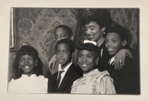 Black and white image of an adult woman with five children. They all smile and she drapes her arms over their shoulders.