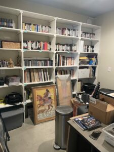 An office with tall bookshelves full of tapes, folders, and other objects. Boxes and framed objects sit on the floor.