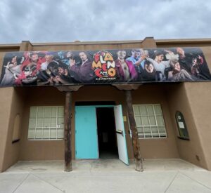 A large banner stretched across a tan building with a bright blue front door. The banner features the faces and gestures of many dancers.