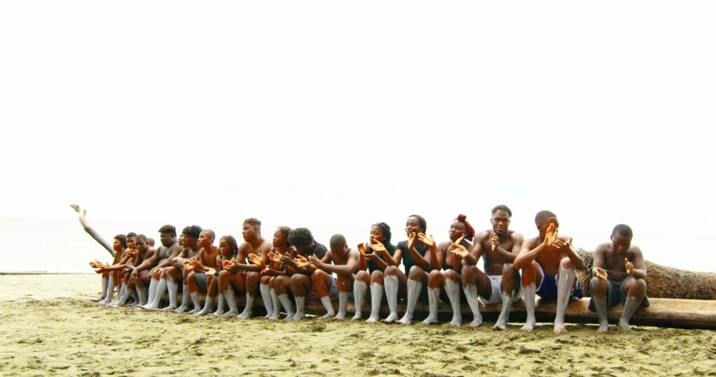 19 Black artists sit on a log on the sand. They hold their hands up like they are clapping.