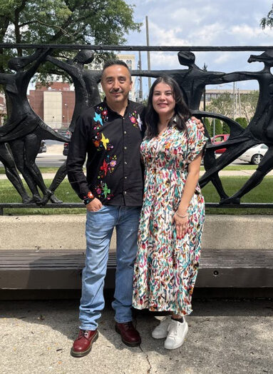 Two smiling people with their arms around each other outdoors in front of a statue depicting dancers.