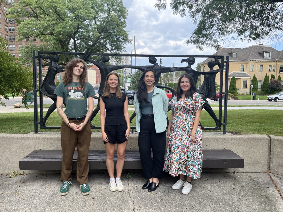 Four smiling individuals standing outside in front of a statue depicting dancers.