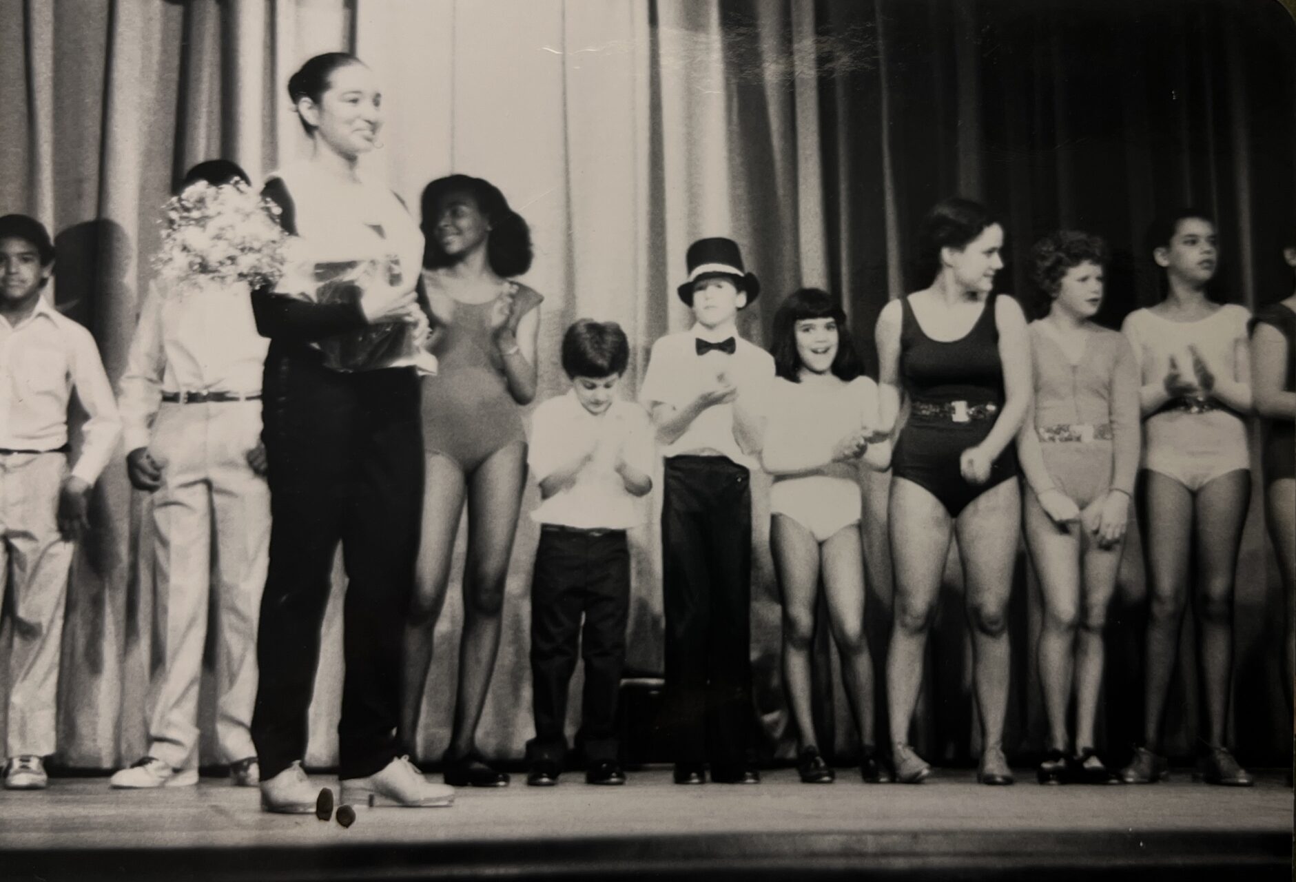 Black and white image of an adult and a long row of dancers and children on stage smiling.