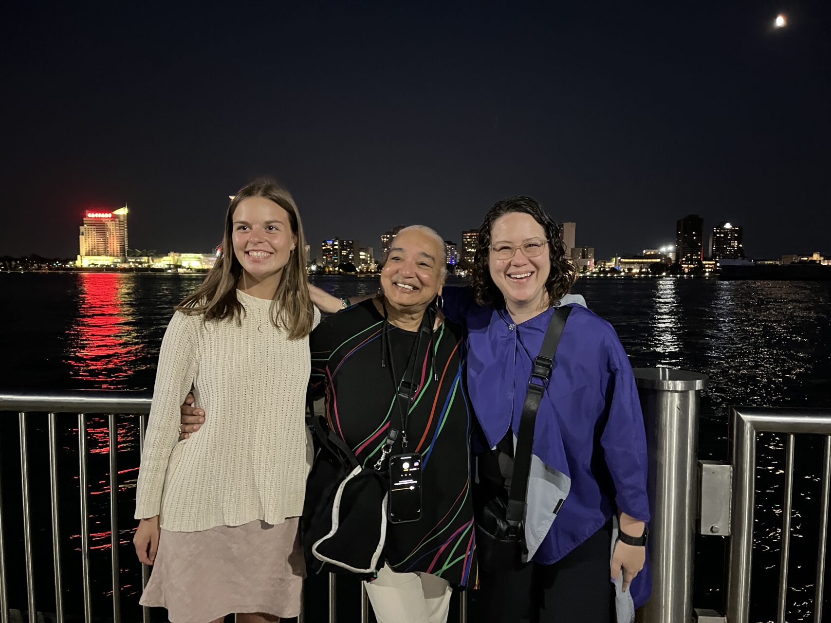 Three people stand smiling with their arms around each other outside at night.