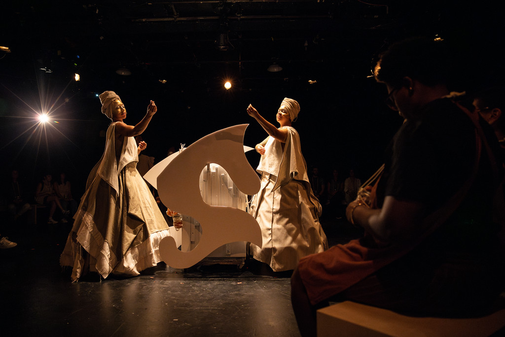 Two artists in large, white, draping gowns and white headdresses gesturing their arms out next to a curvy, irregularly shaped white object.