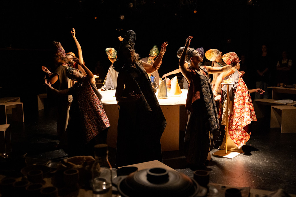 Ten dimly lit artists in elaborate, colorful draping gowns and headdresses surrounding a table covered in handcrafted vessels and gesturing their arms.