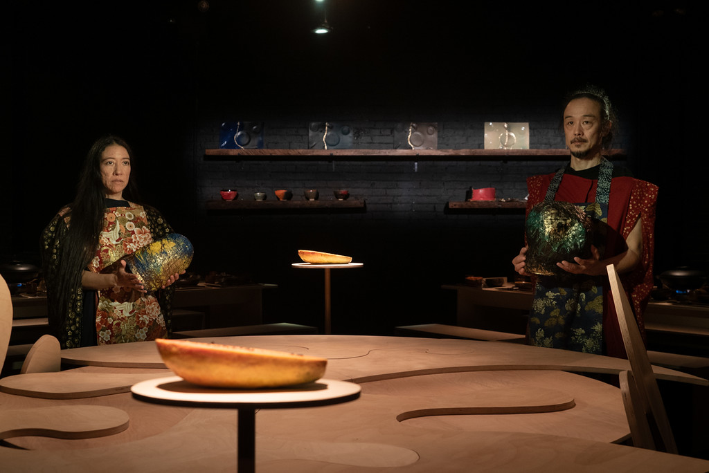 Two artists stand behind an irregularly shaped table holding handmade vessels. A sliced papayas sits on either side of them, and the back wall behind them has more handmade objects on shelves.