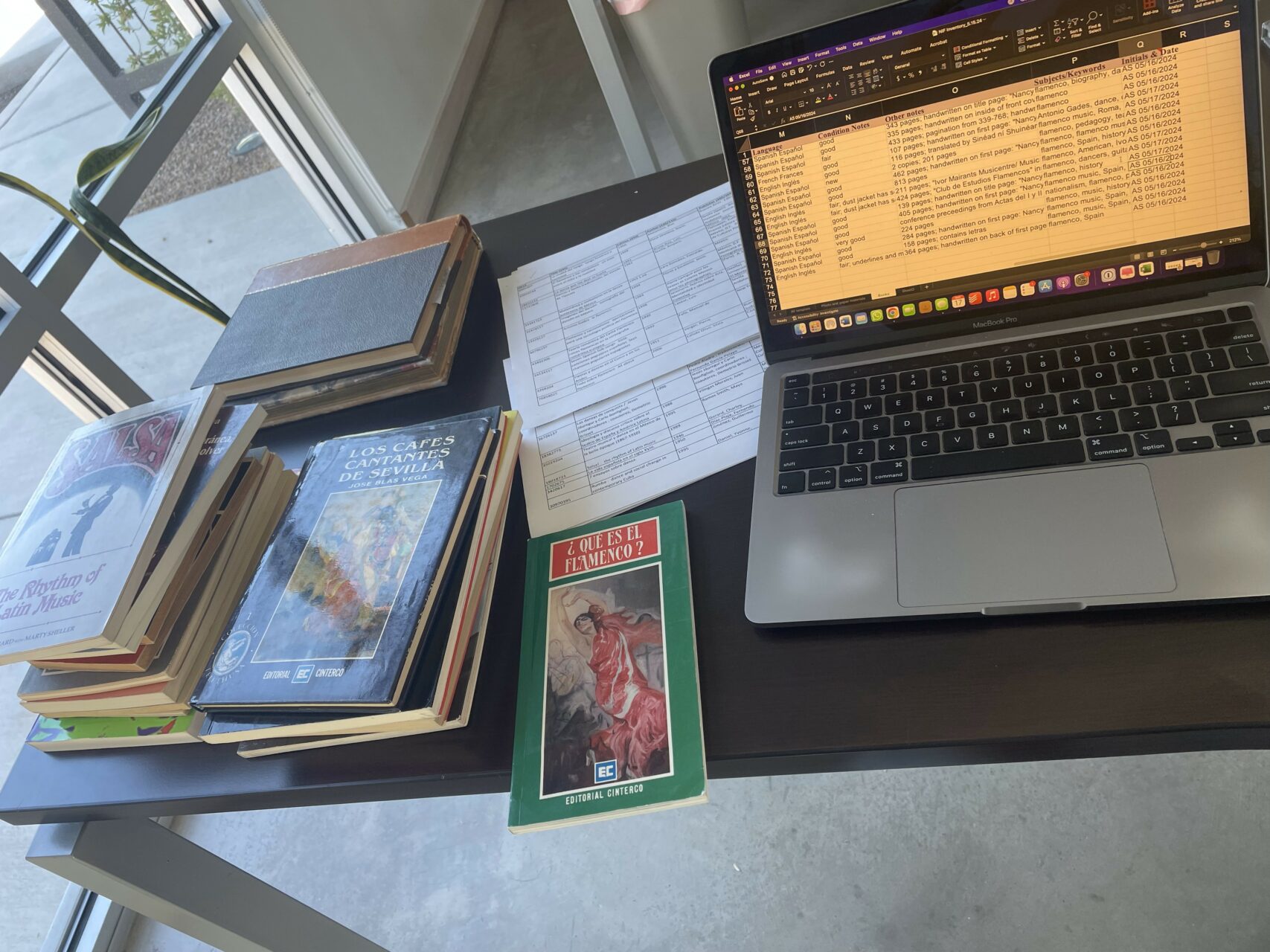 A table with a laptop open to an excel sheet and stacks of books and papers.