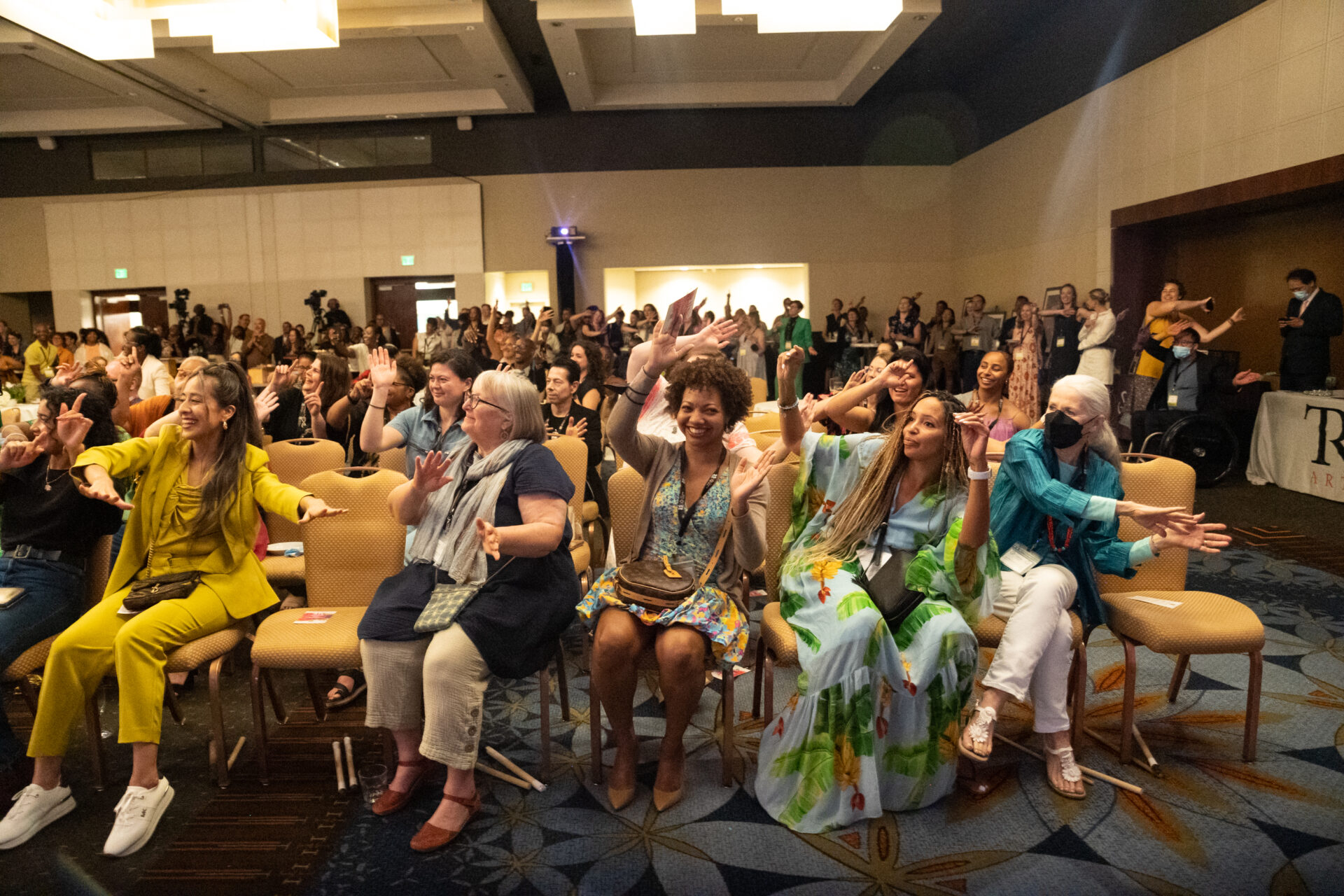 Photo of a crowd of seated dance professionals waving their arms above their heads and smiling at a Dance/USA convening