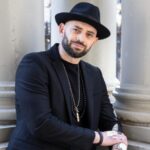 Christopher leans back on his left elbow and grasps his left wrist with his right hand. He wears a full black suit, silver jewelry, and a black fedora hat. There are three columns of classical architecture behind him. He smiles shyly as he looks at the camera. Photo by Walter Wlodarczyk.