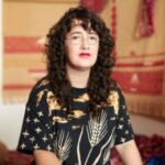 iele paloumpis, a white genderqueer person with dark curls and eyes, looks directly and confidently at the camera. They wear square gold-rimmed glasses, a bold, two-toned lip in red and hot pink, and a shirt emblazoned with images of clouds and wheat, created by Indigenous-American designer Noel Bennetto. In the background, iele’s textile studio displays weavings and embroidered tapestries in warm, rich tones. Photo by Lily Olsen.