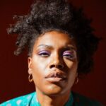 Jenn Freeman | Po’Chop is a Black queer woman with an afro. Her black hair is styled asymmetrically with curls spiraling down both sides of her face. Her eyes are adorned with purple eye shadow. She is wearing gold hoops and a turquoise button up shirt that features a red art deco pattern. A ray of light illuminates her face and shoulders while a shadow streaks across her forehead and right shoulder. Photo by Anjali Pinto.