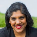Image description: Bhumi B Patel, a queer, brown skinned, South Asian Gujarati, femme person with black and gray hair sits against a field of grass blurred in the background. She wears a v-neck black jumpsuit and bright red lipstick. She has brown eyes and a silver hoop nose ring. Photo by Lara Kaur.