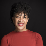 A medium light-skinned woman wearing a deep rustic orange colored short sleeve top in front of a charcoal gray backdrop. She is smiling with red lipstick and her hair is styled in a medium length naturally curly fro-hawk. Her arms are crossed in front and her head is slightly tilted to the right. She is wearing small dangle pearl earrings. Photo by Melisa Cardona.