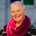 Petra Kuppers, a white queer cis woman of size, head tilted, smiling with twinkling eyes, with yellow glasses, shaved head, pink lipstick, purple scarf, polka-dot top, a hand caressing the handlebars of Scootie, her mobility scooter, before an urban building with colored glass windows. Photo by Tamara Wade.