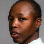 A headshot of a Black person of African descent with brown skin, very low hair cut, and black square earrings in their ear. Her upper body is in profile, and head is looking towards the camera. They are wearing a sweatshirt with aquamarine trimming, followed by white on the neck collar, and aquamarine color blocking on the top half of the shirt, that has quilt-like stitching. Photo by Maria J. Hackett.