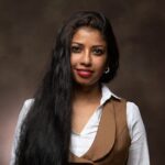 A dark brown skinned woman, long black hair, wearing a white shirt, khaki waistcoat and skirt, with a nose ring on the right side of her nose per the Tamil tradition, smiles. Photo by Nathan Cornetet/Fusion Photography.