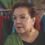 Gema Sandoval looks into the distance wearing a green and yellow rebozo over her black top. She is wearing red and gold filigree earrings and is standing in front of a rack full of colorful costumes. Photo by Juan Escobedo.
