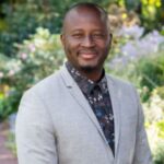 A black man from Burkina Faso stands smiling. He has a bald head and wears a silver suit and a dark flowery shirt while standing in front of trees and bush of colorful flowers. Photo by David Crow.