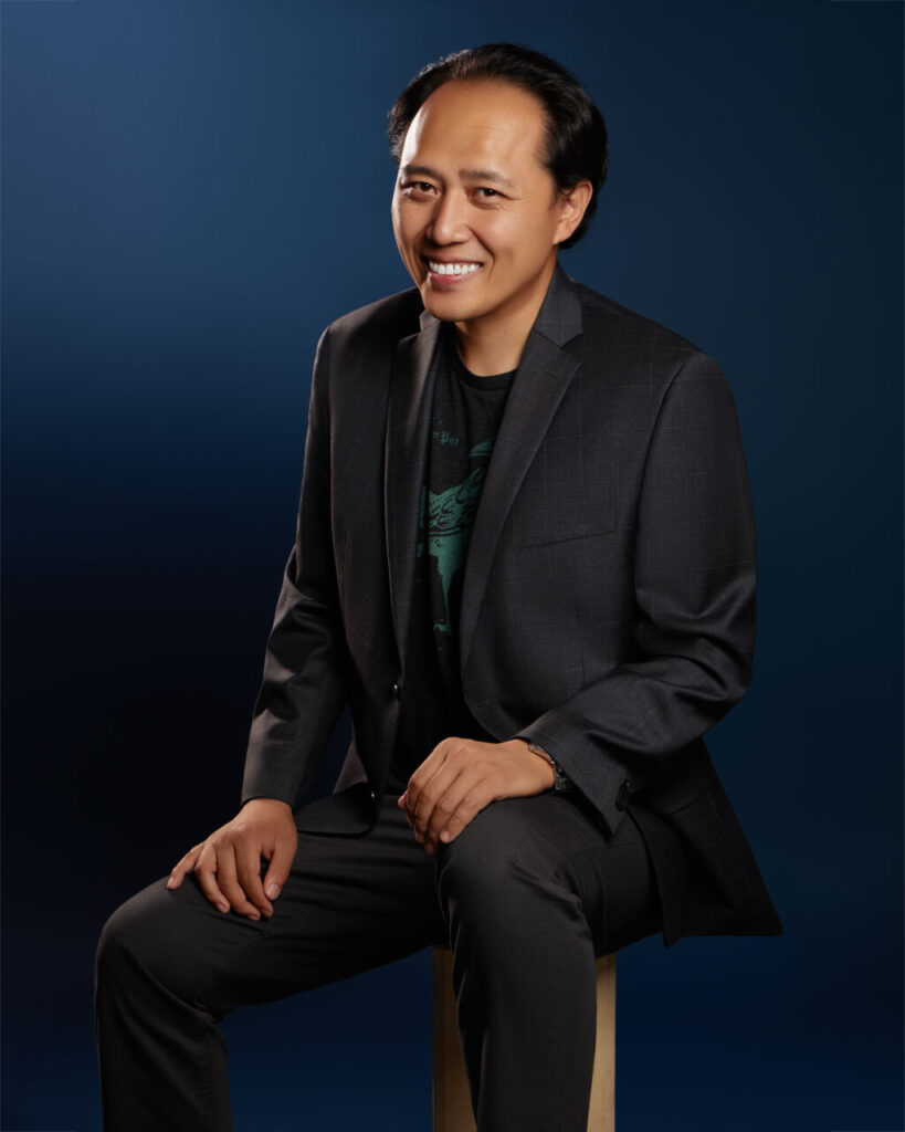 Haowen is a Taiwanese man with dark, wavy hair, warm brown eyes, and a light-medium complexion. Wearing a charcoal suit over a dark shirt, he sits on a stool with relaxed posture, hands resting naturally. He smiles openly toward the camera against a smooth deep-blue backdrop.