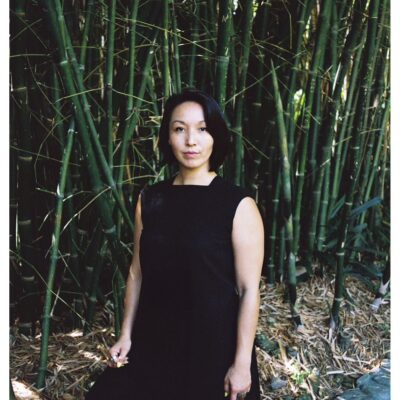 A mixed East Asian woman with short, black hair wears a black dress and stands strong among a cluster of green bamboo shoots that stretch overhead
