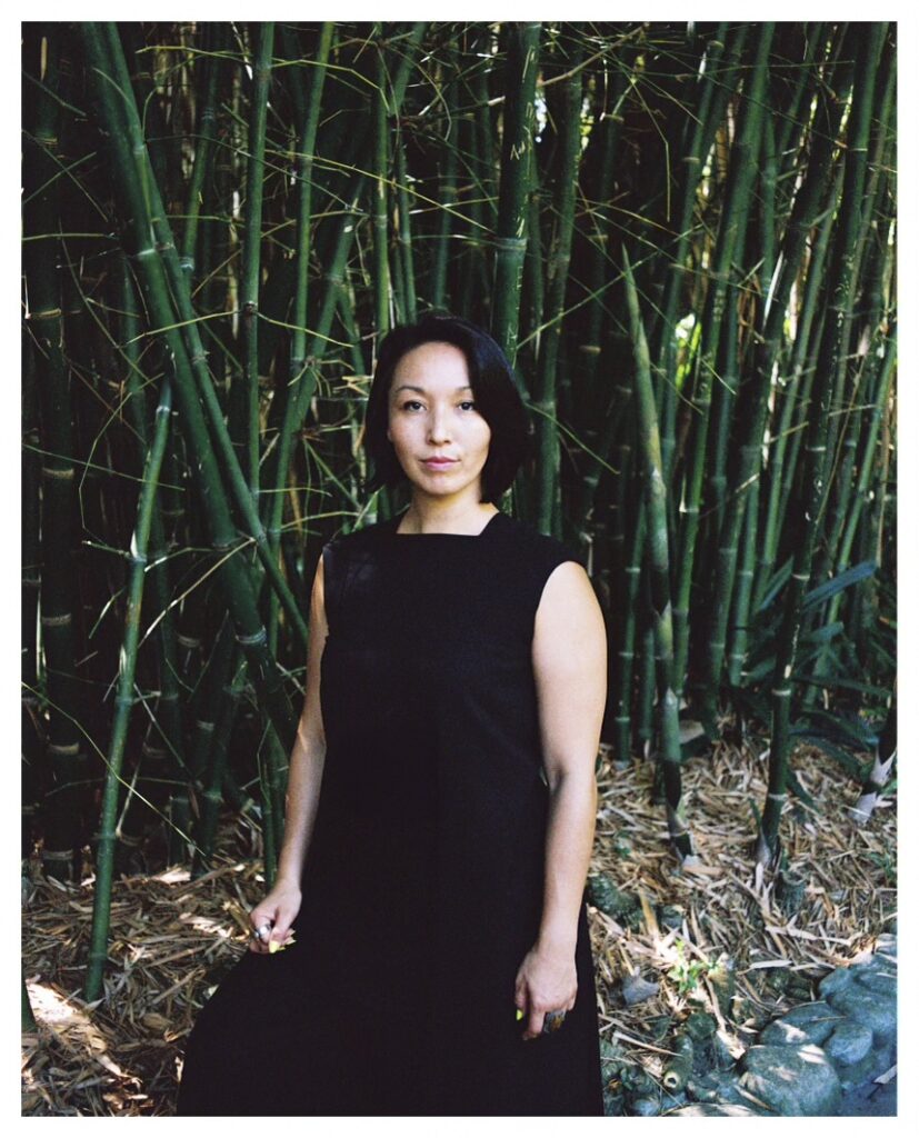 A mixed East Asian woman with short, black hair wears a black dress and stands strong among a cluster of green bamboo shoots that stretch overhead